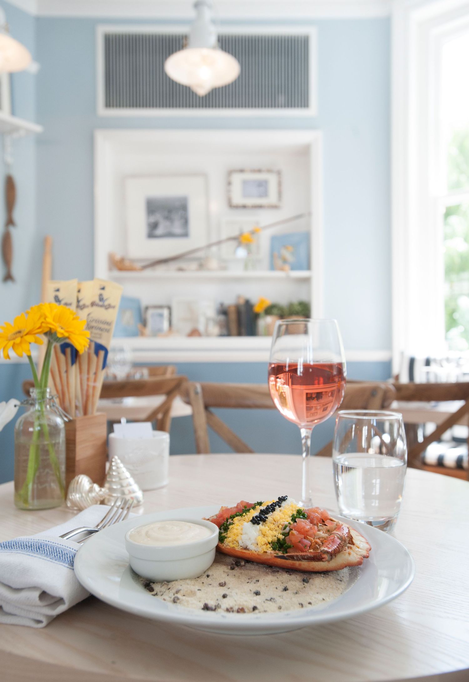 Elegant restaurant table with smoked salmon toast, rose wine, and yellow flowers in a charming blue and white coastal dining room.