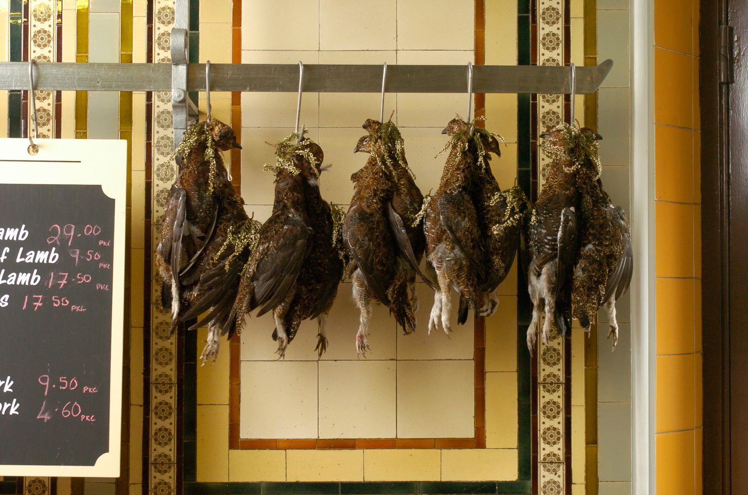 Dead game birds hanging on a rack in a traditional butcher shop with a price board visible.