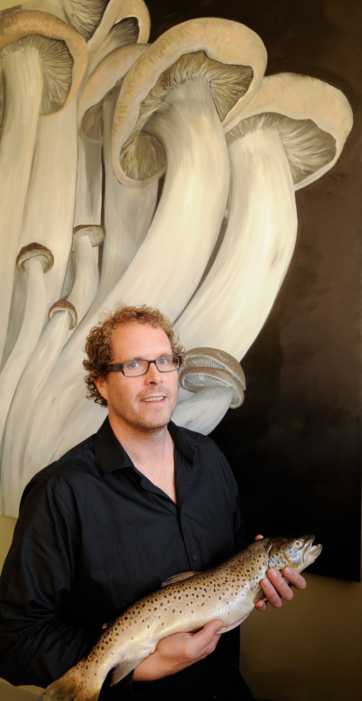 Man in black shirt holding a fresh trout fish, standing before a large mushroom painting artwork.