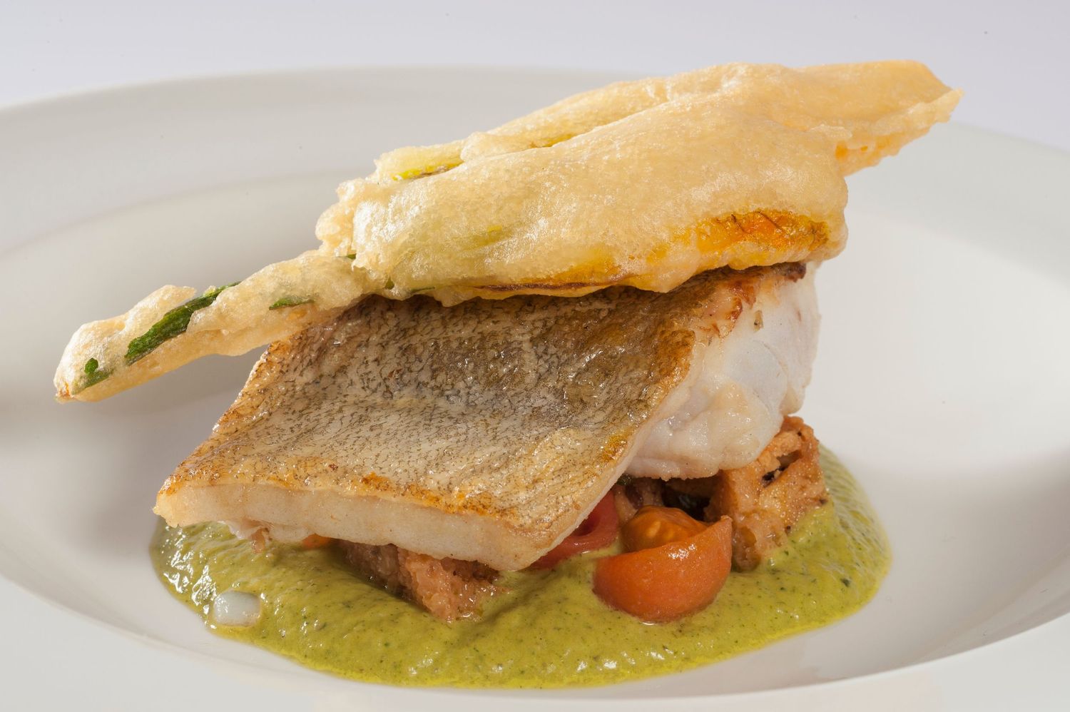 Elegant plated fish dish with crispy skin, green herb sauce, cherry tomatoes, and a golden tempura crisp on white plate.