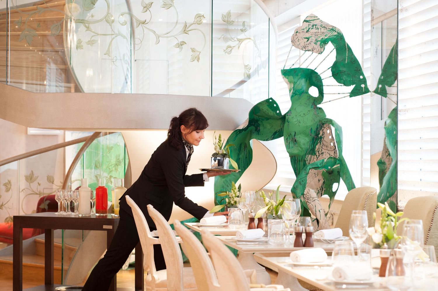 Waitress setting tables in an elegant restaurant with green artistic wall murals and white chairs.