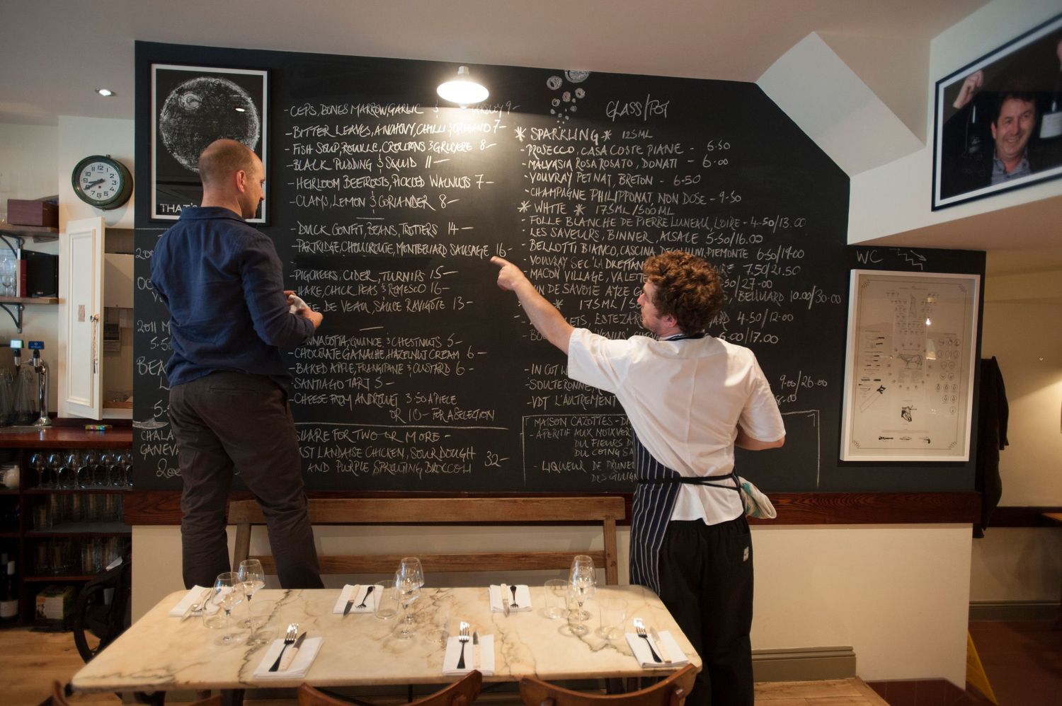 Restaurant staff writing daily specials on a large chalkboard menu above a neatly set dining table.