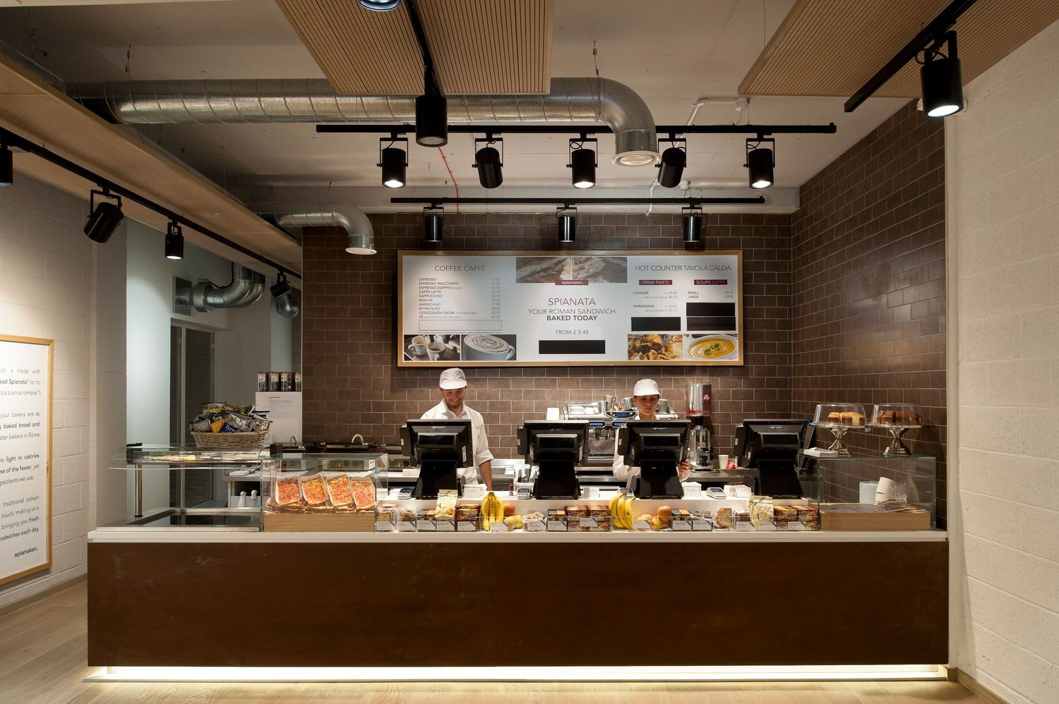 Modern bakery cafe counter with display cases, espresso machines, and illuminated menu boards in a warm wood interior.