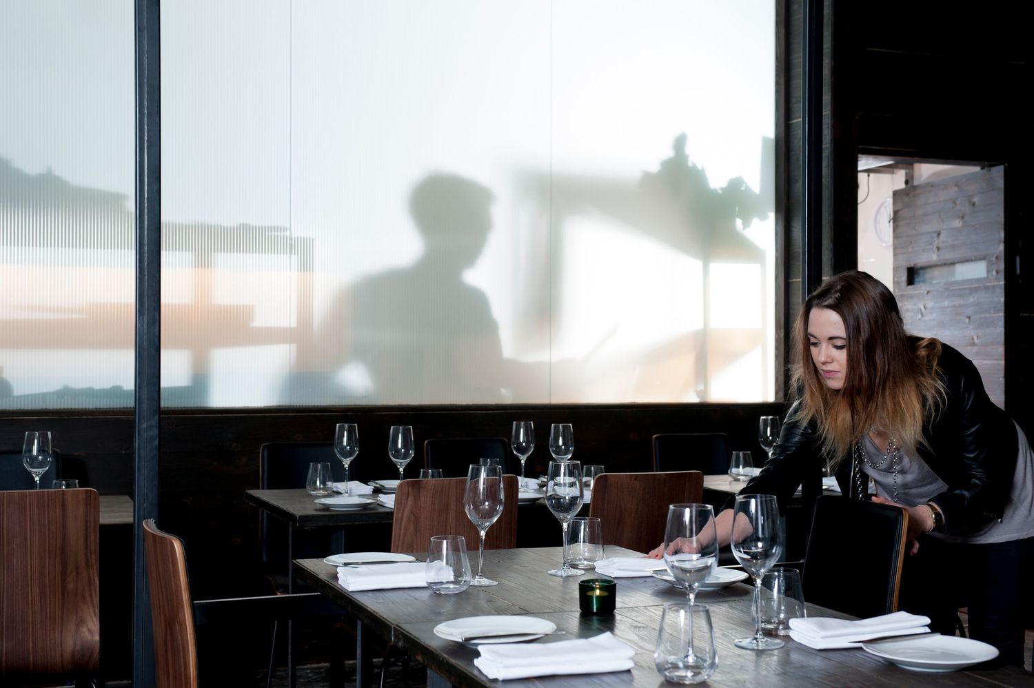 Woman setting a restaurant table with wine glasses near a large window with shadows visible.