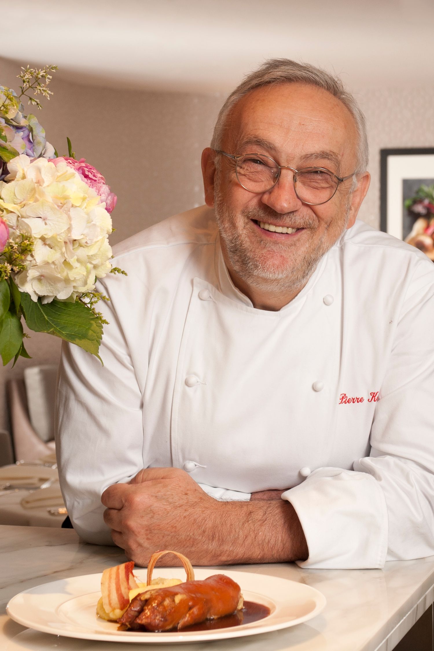 Smiling chef in white uniform poses with a plated dish beside a floral arrangement in a restaurant setting.