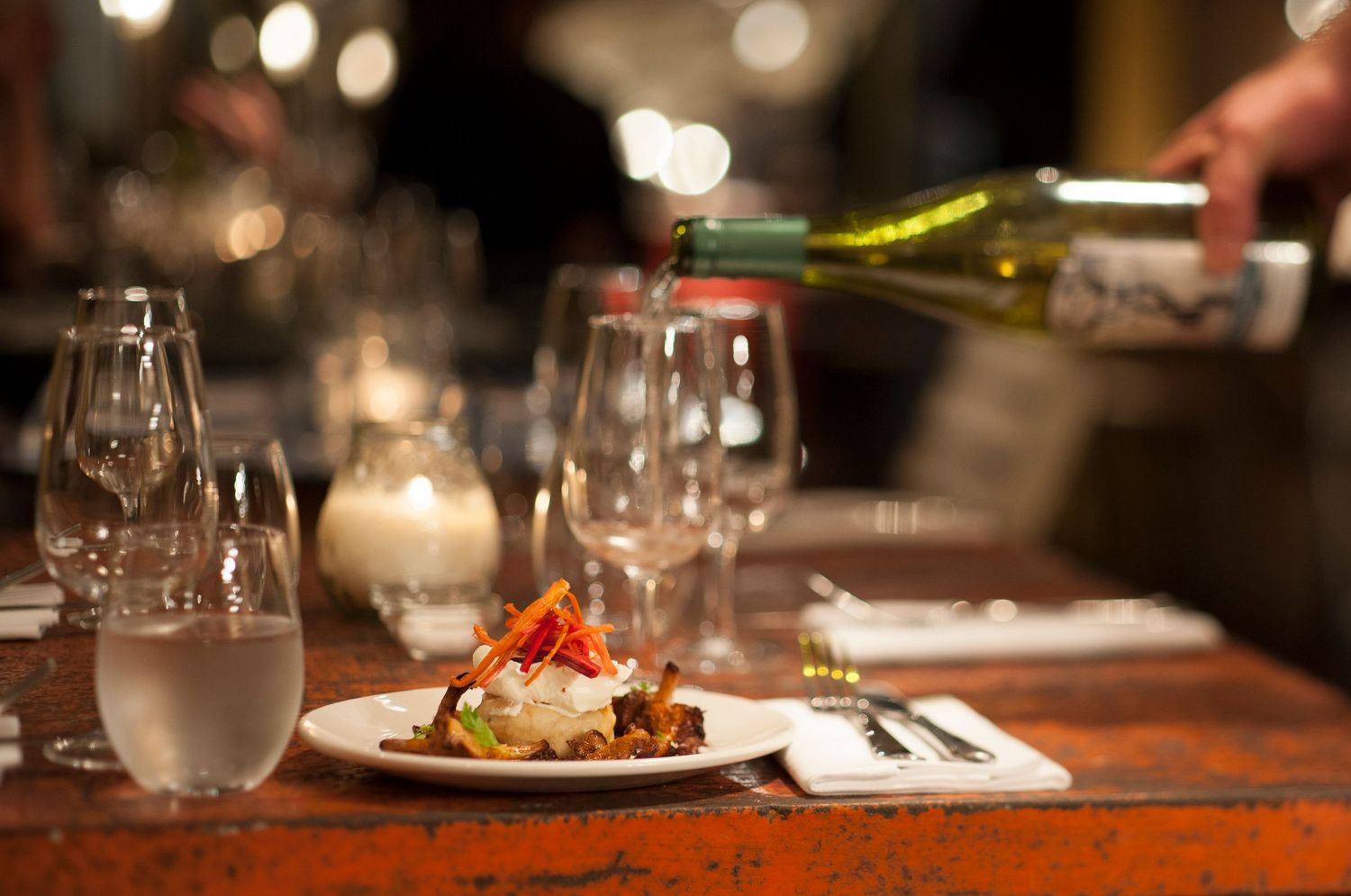 Elegant restaurant table with a gourmet dish, wine glasses, and a bottle of white wine being poured in dim lighting.
