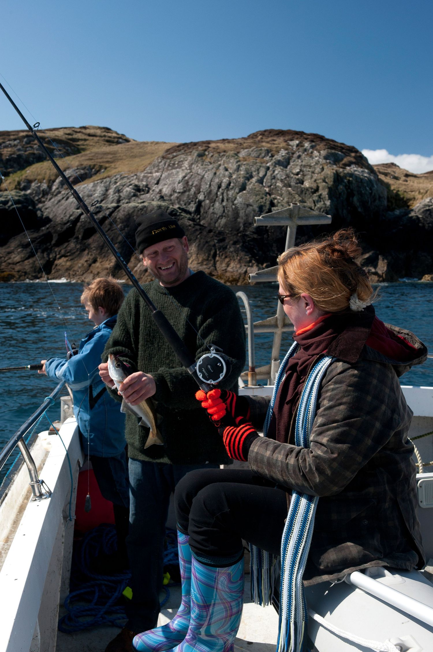 Family fishing on a boat near rocky Scottish coastline on a sunny day, with man holding a caught fish.