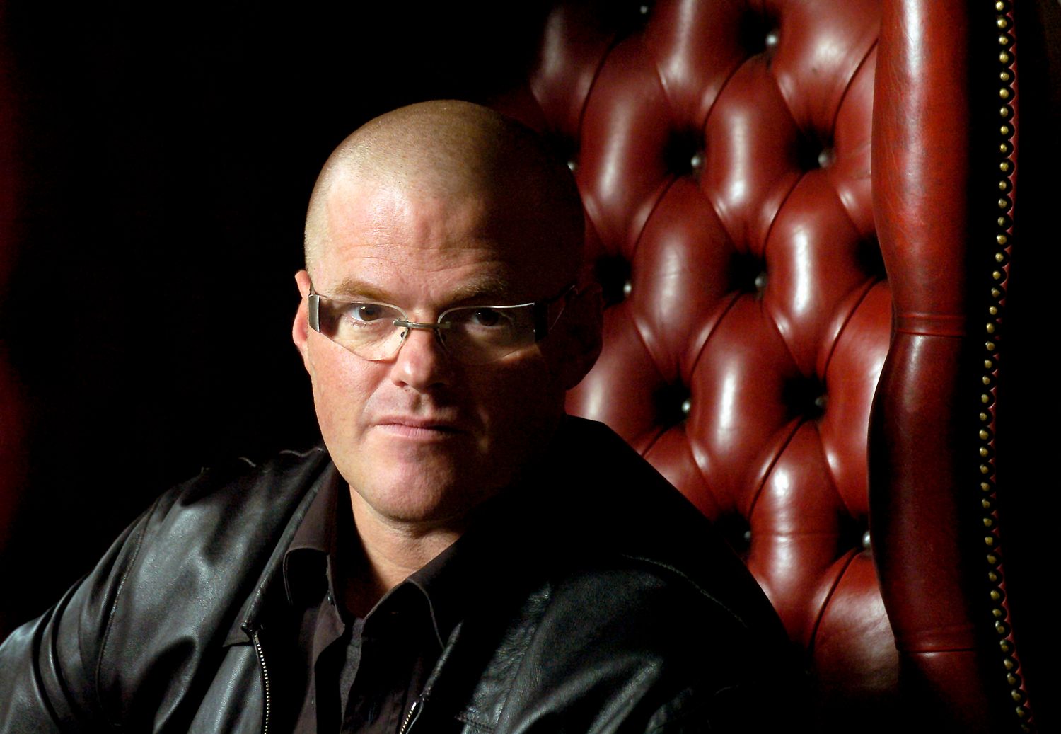 Bald man with glasses sitting in a tufted leather chair, wearing a black jacket, looking directly at camera.
