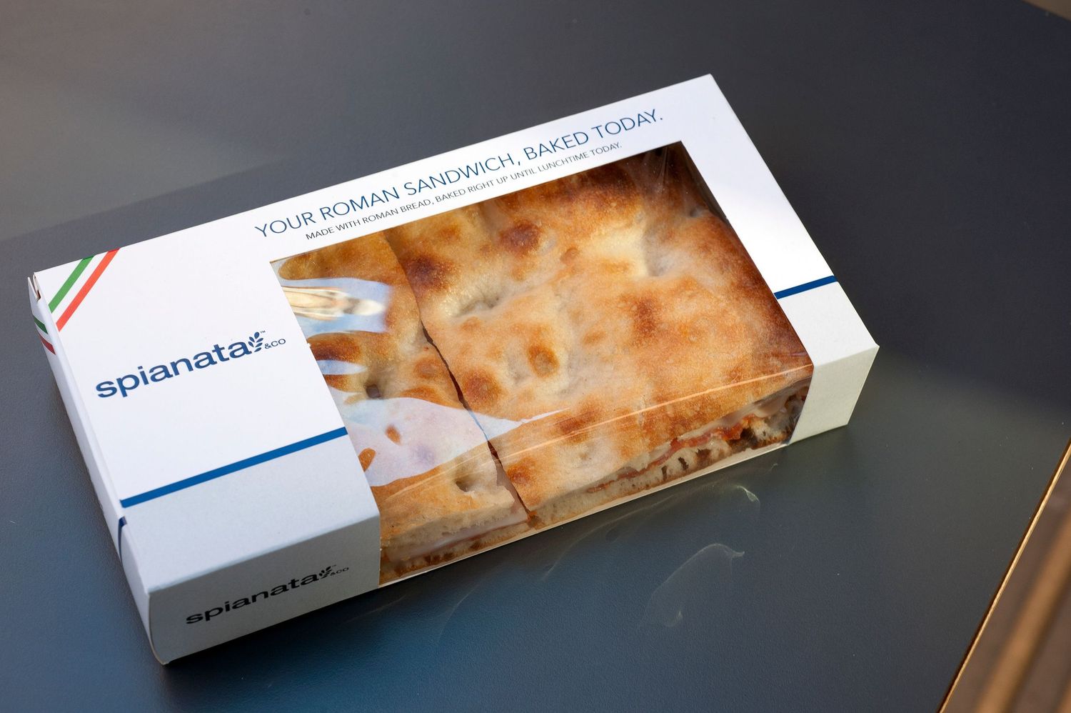 Spianata Roman sandwich in branded packaging, freshly baked today, displayed on a gray surface.