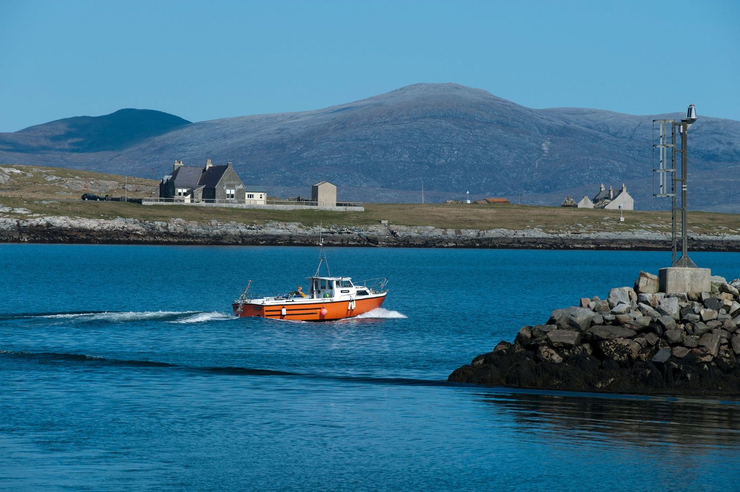 Orange boat speeding through calm blue waters near a rocky harbor with Scottish highland village and mountains in background.