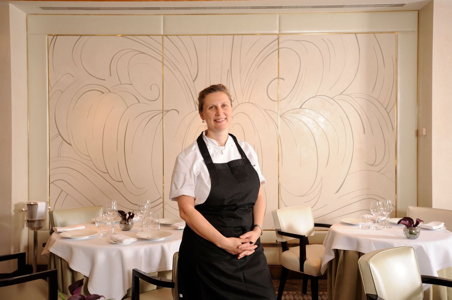 A smiling female chef in a black apron stands in an elegant white fine dining restaurant with decorative wall panels.