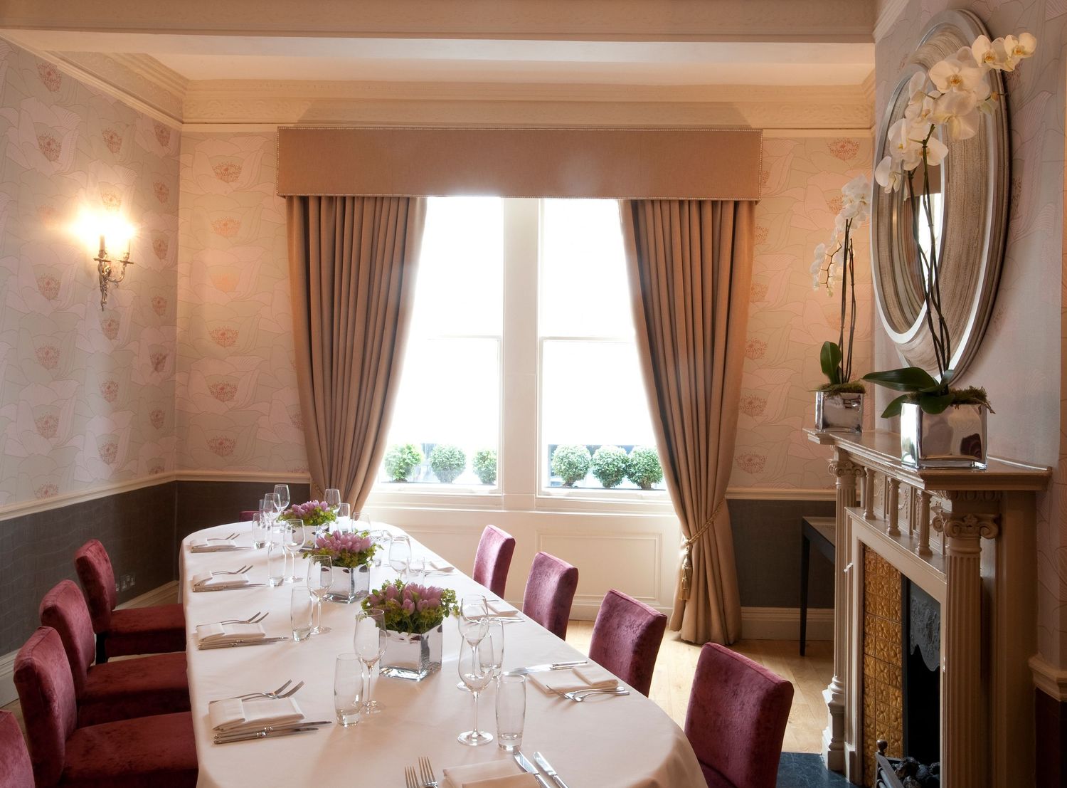 Elegant private dining room with burgundy chairs, long white-clothed table, fireplace, and draped windows in soft neutral tones.