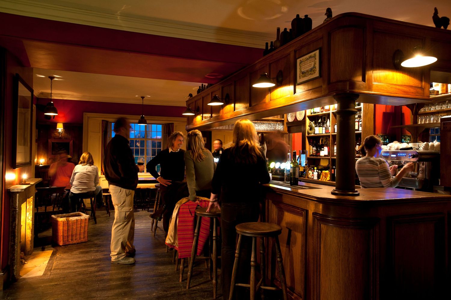 Cozy dimly lit pub bar with patrons gathered around a wooden counter, warm amber lighting, and bar stools in a classic tavern setting.