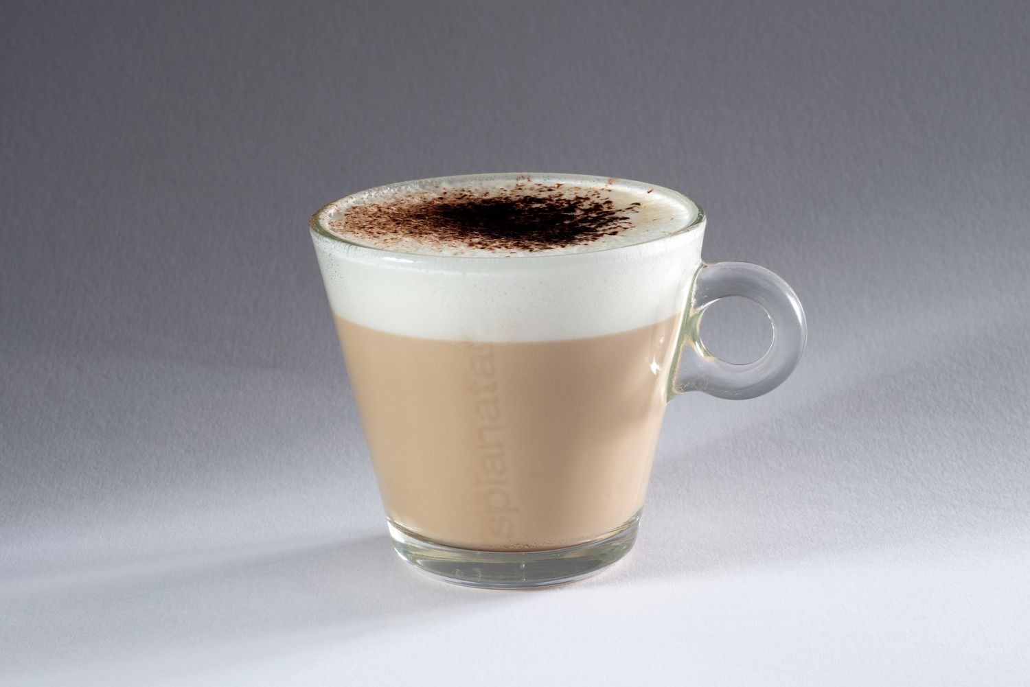 Glass cup of layered cappuccino with frothy milk foam and cocoa powder on top, set against a grey background.