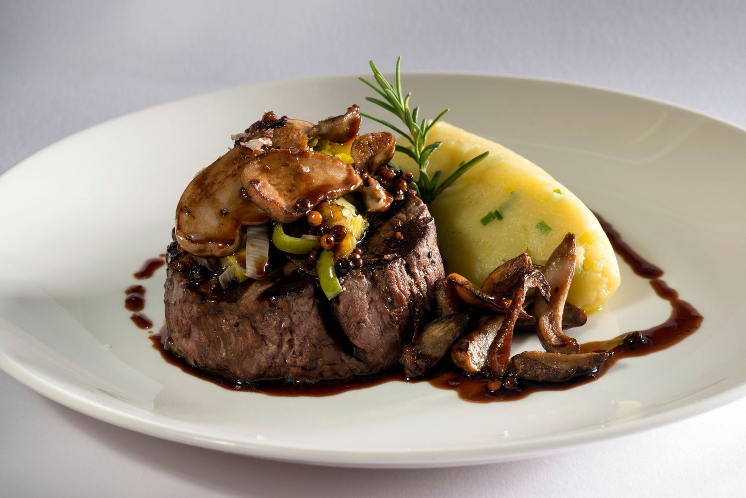 Elegant plated beef filet with mushrooms, caramelized onions, creamy mashed potatoes, and rich red wine sauce on white plate.
