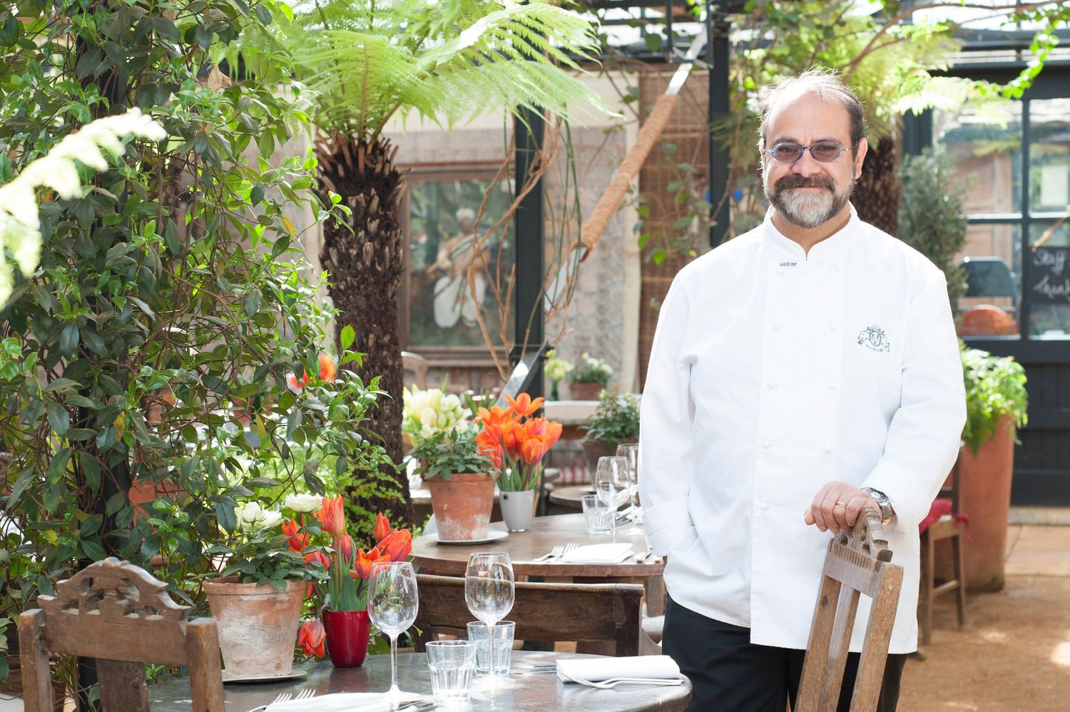 A smiling chef in white uniform stands among lush greenery beside an elegantly set restaurant table.