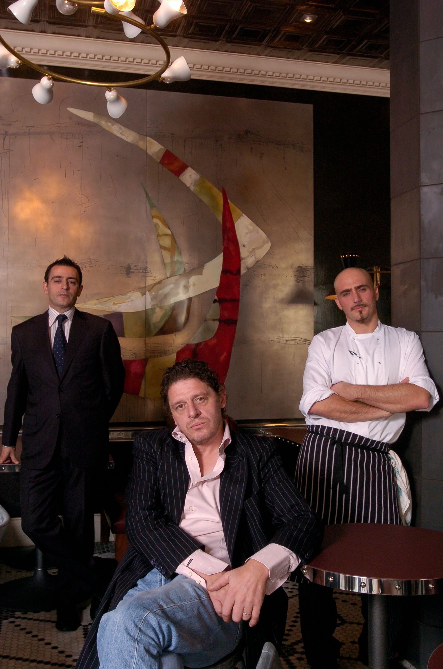 Three men pose in an upscale restaurant, one seated in a suit, one in chef whites, before a large abstract painting.