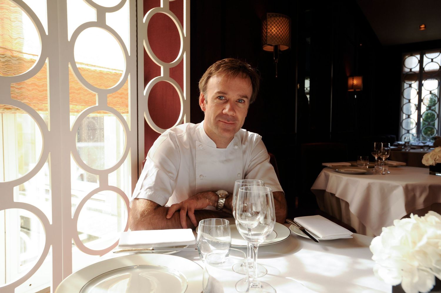 A chef in white uniform sits at an elegantly set restaurant table with wine glasses and white linen.