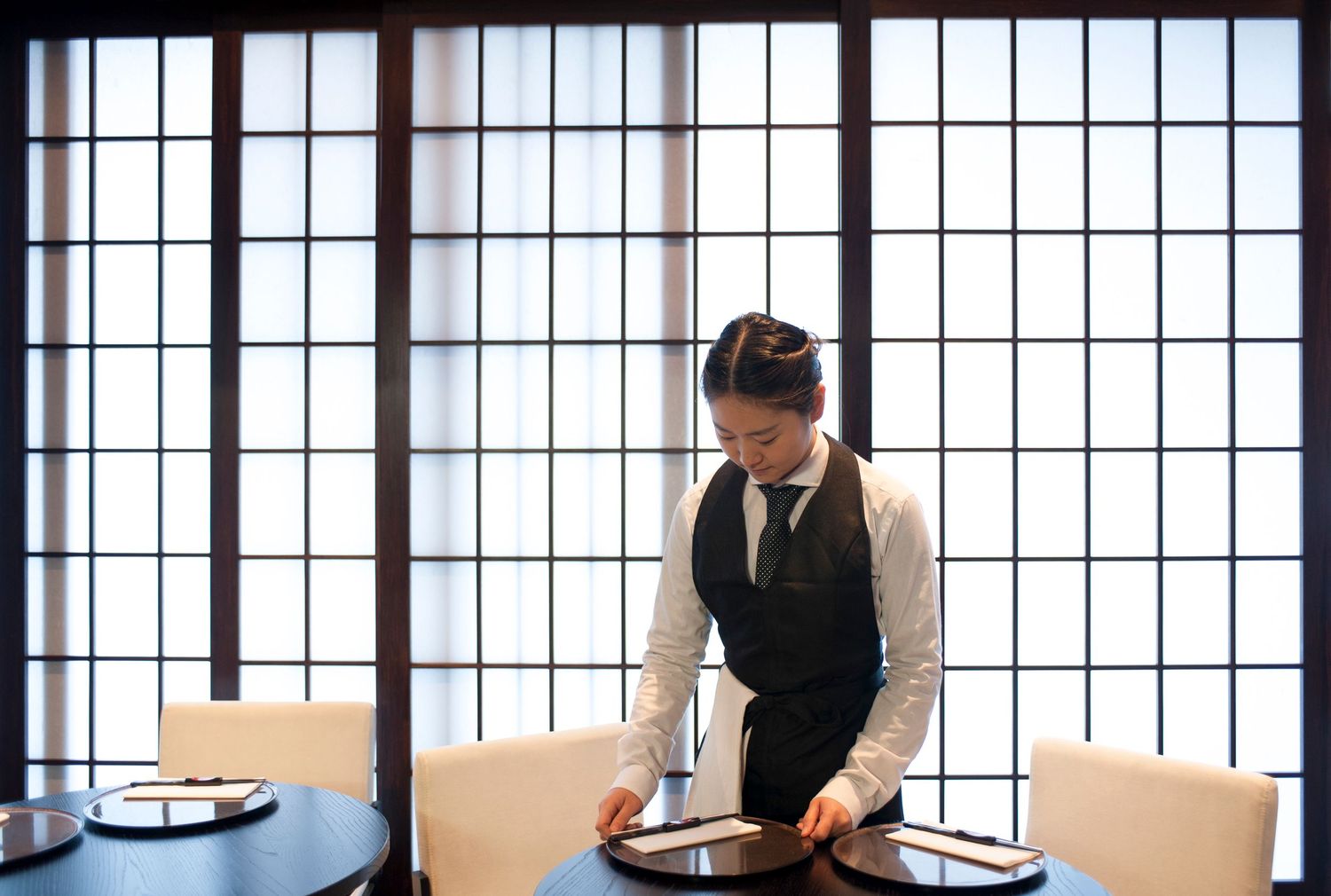 A waitress in formal attire carefully sets a round table in an elegant Japanese restaurant with shoji screen windows.