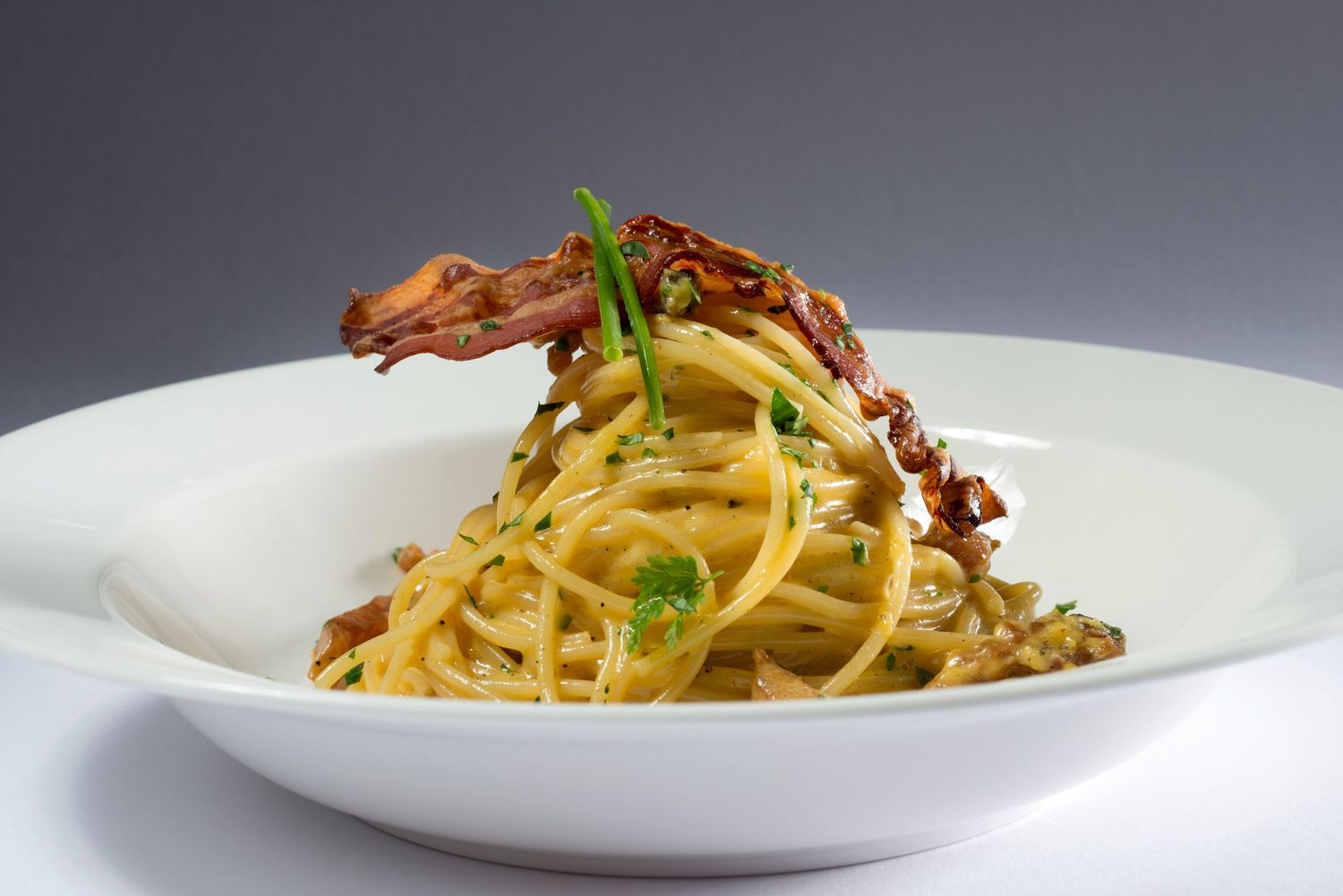 A plate of spaghetti carbonara topped with crispy bacon and fresh herbs served in a white bowl.