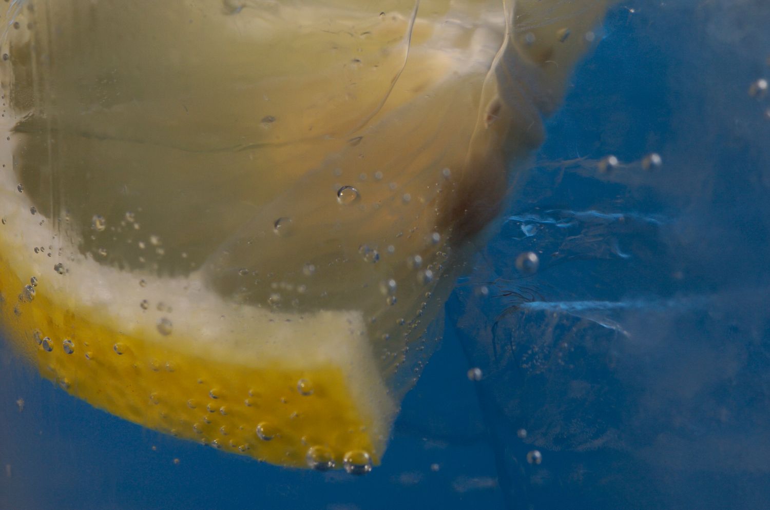 Close-up of a lemon slice submerged in sparkling water with bubbles against a blue background.