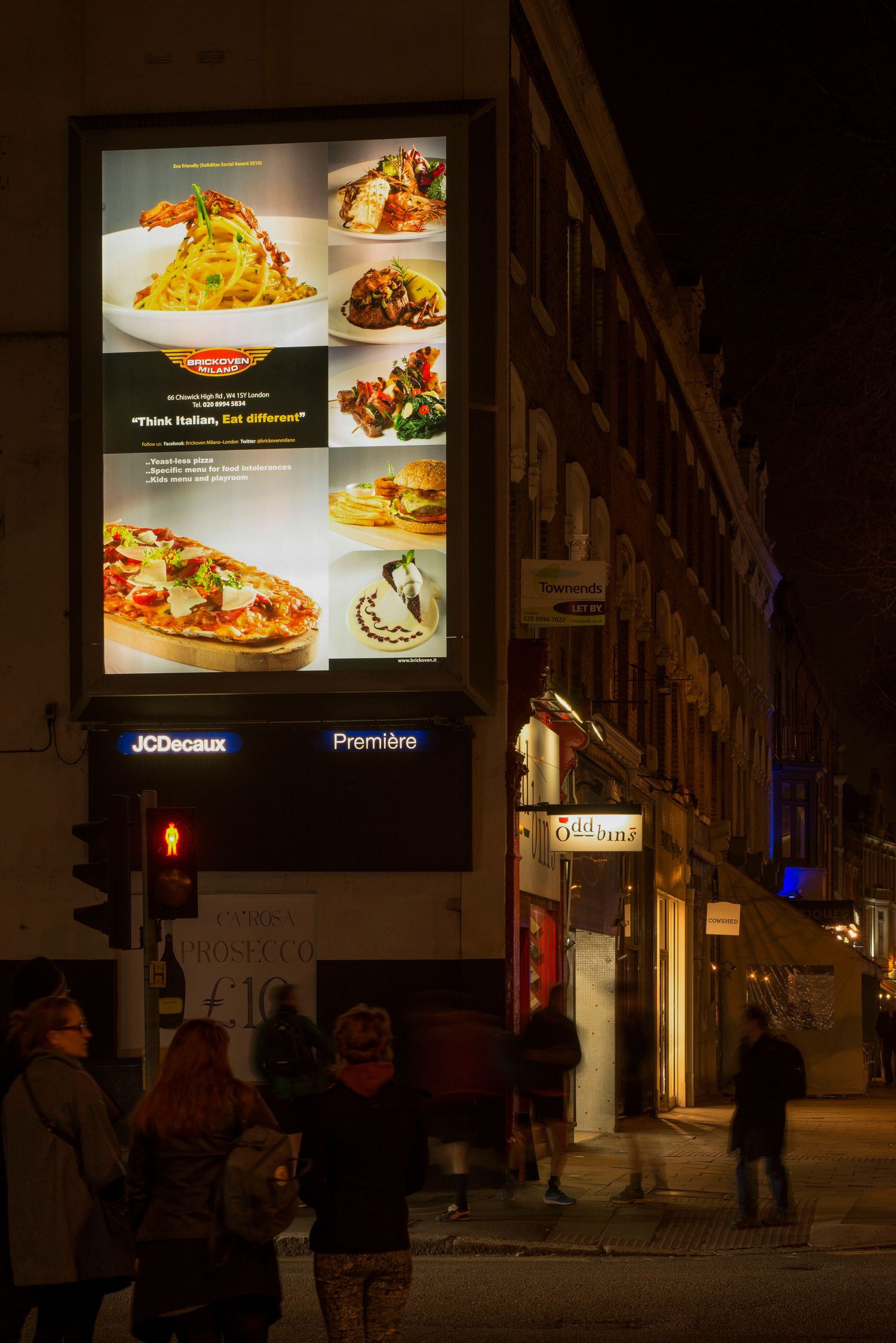 Illuminated JCDecaux food advertisement billboard on a busy night street corner with pedestrians walking below.