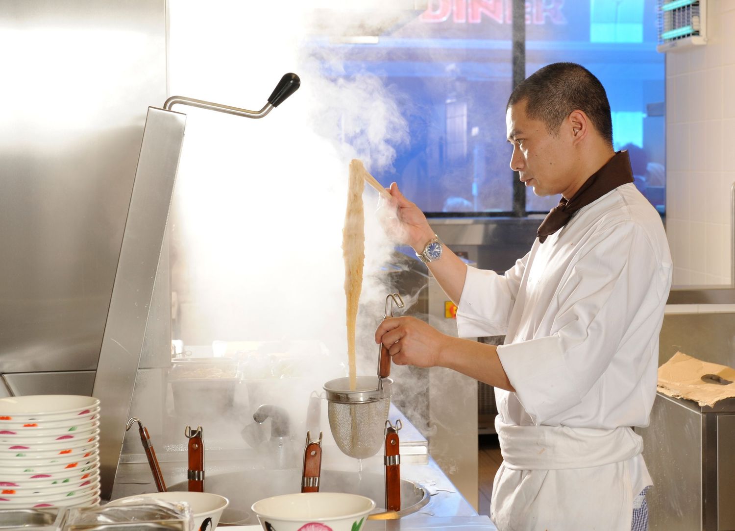 A chef in white uniform cooking with steam rising in a professional kitchen with bowls and bottles nearby.