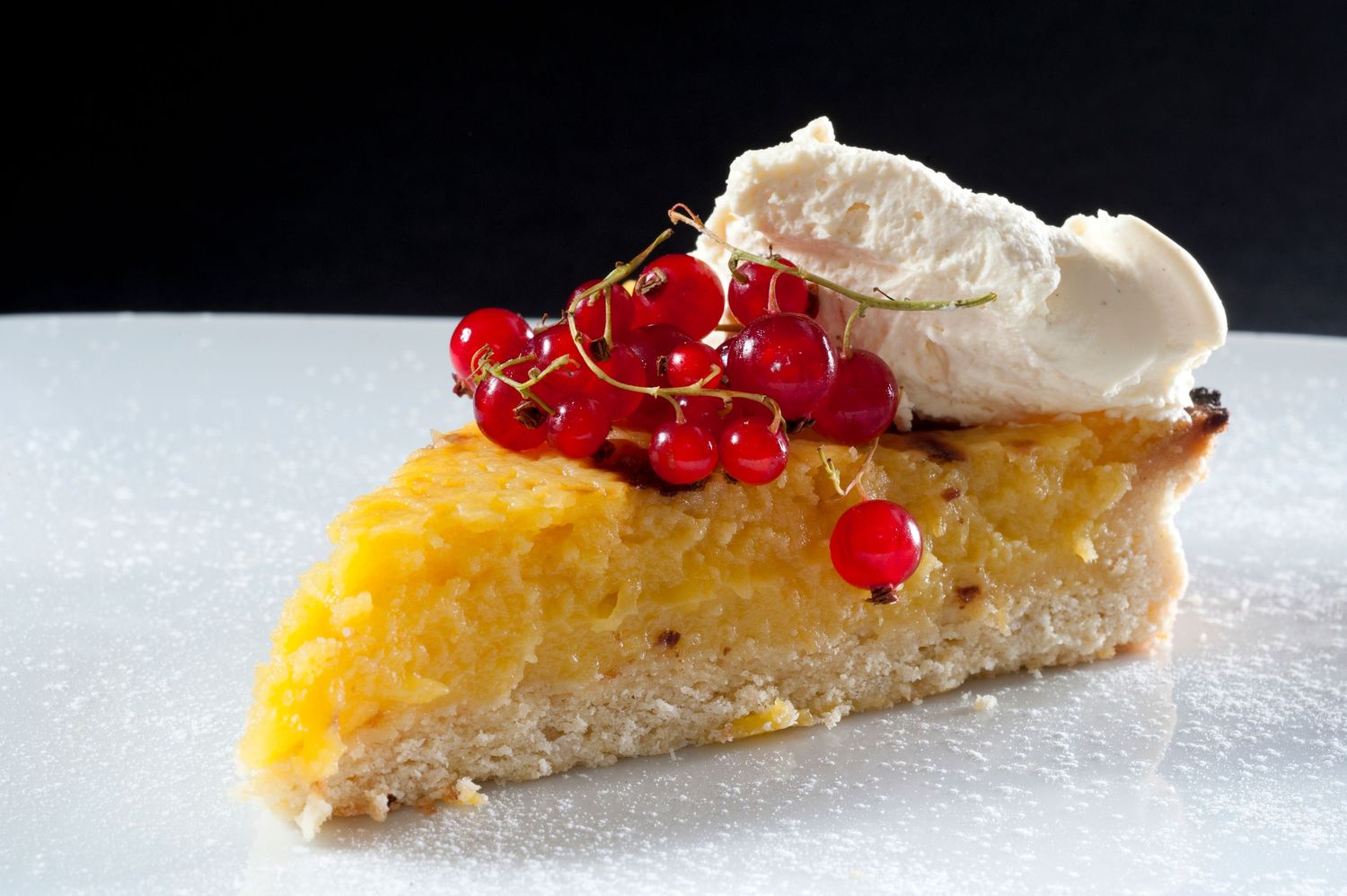 A slice of golden tart topped with fresh red currants and whipped cream on a white plate.