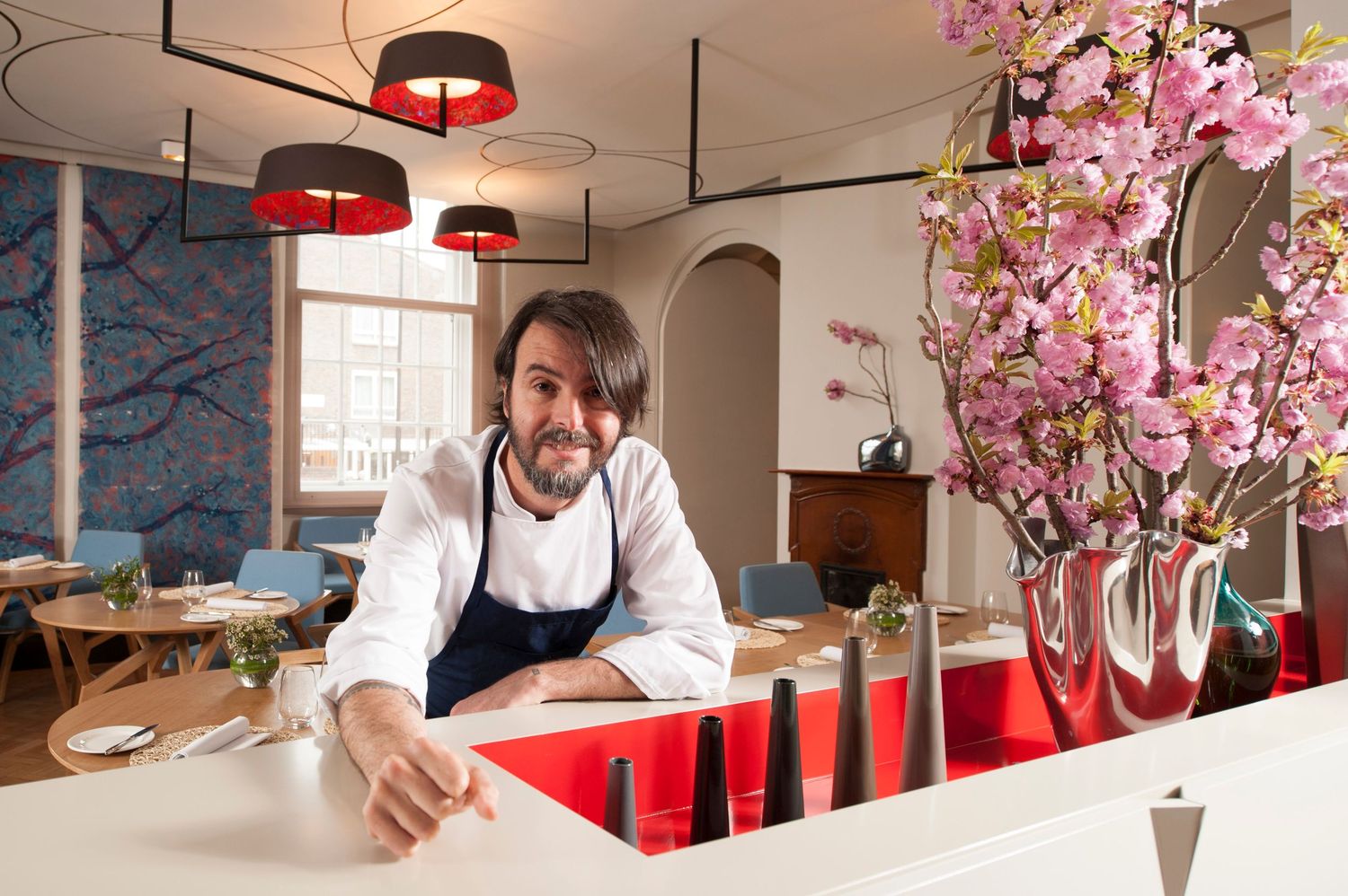Chef in white apron smiling at restaurant counter with pink cherry blossoms and red pendant lights.