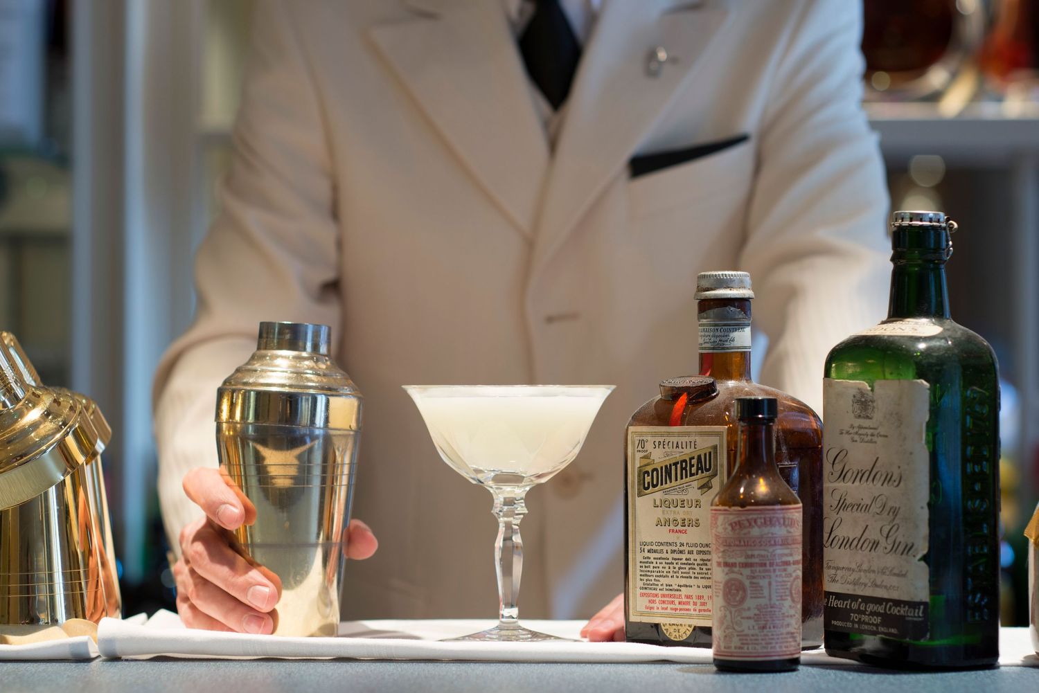 Bartender in white jacket preparing a cocktail with shaker, coupe glass, and vintage spirit bottles on bar.