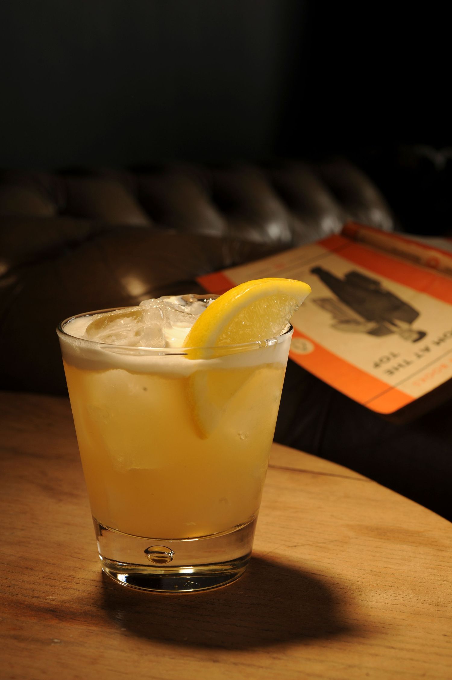 A whiskey sour cocktail with ice and lemon wedge garnish in a rocks glass on a wooden bar table.