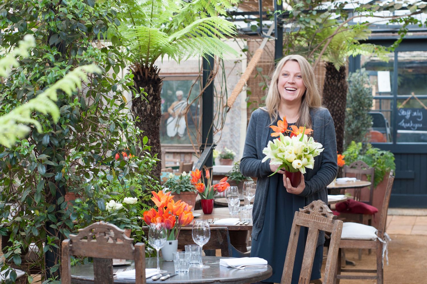 Smiling woman holding white flowers at an outdoor garden dining table surrounded by lush greenery and colorful floral arrangements.