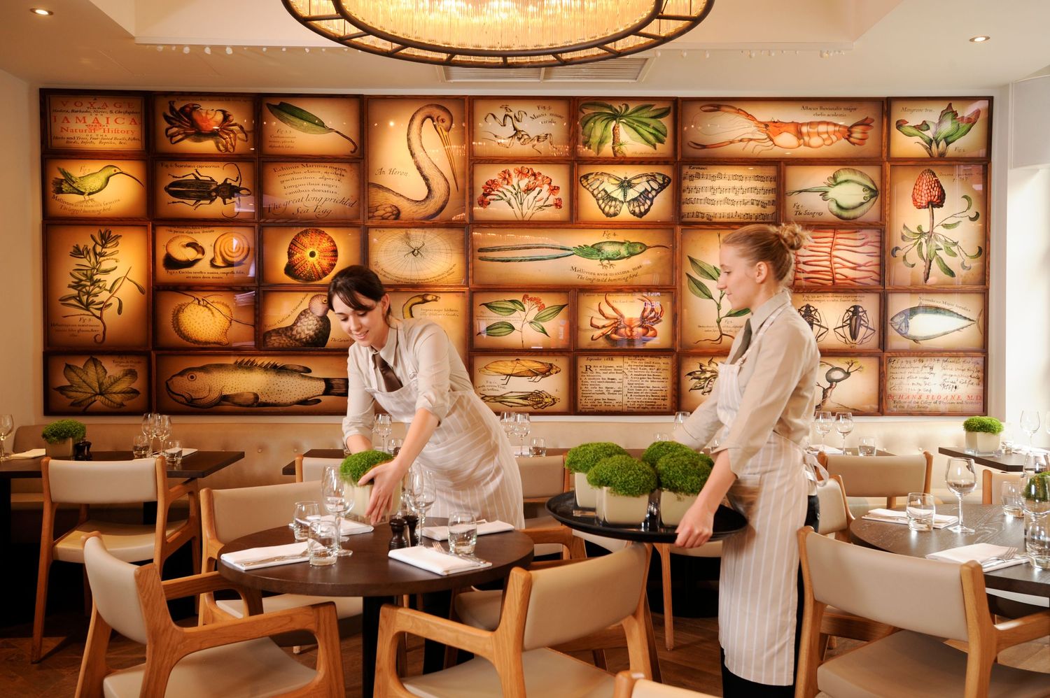 Two servers set elegant tables in a restaurant featuring a decorative wall mural of colorful seafood illustrations.