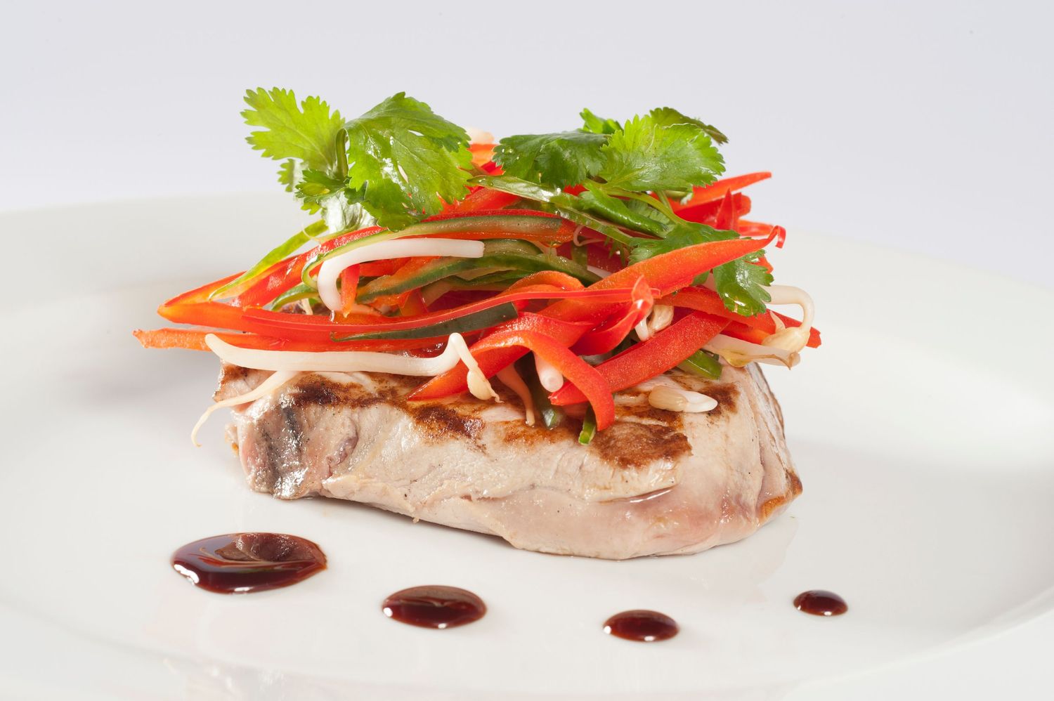 Grilled tuna steak topped with fresh cilantro and julienned red peppers on a white plate with balsamic drizzle.
