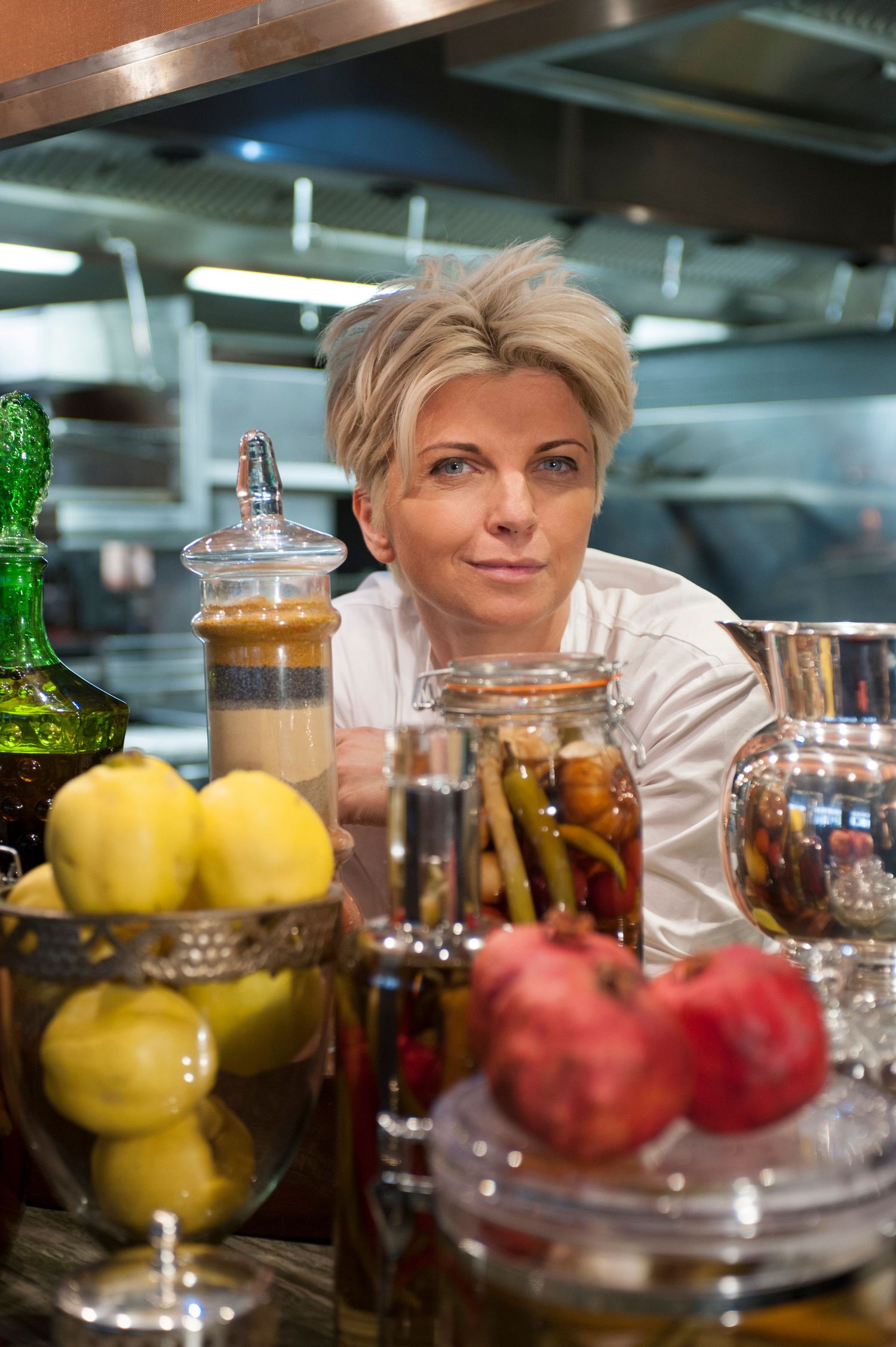 Female chef with short blonde hair poses behind jars of preserved vegetables and fresh fruit in a professional kitchen.