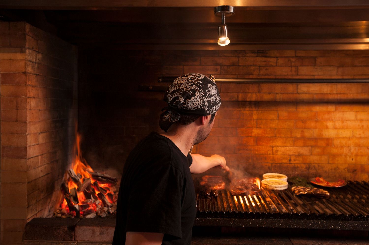 Chef grilling meat over open wood fire in a rustic brick fireplace kitchen.