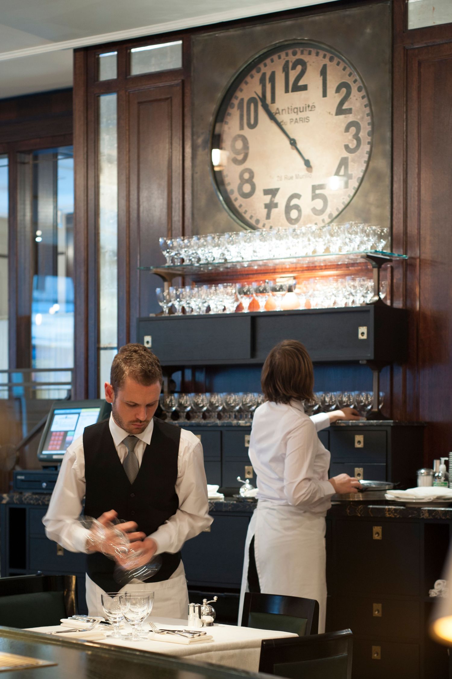 Elegant restaurant staff preparing tables beneath a large vintage clock with wine glasses displayed on shelves in the background.