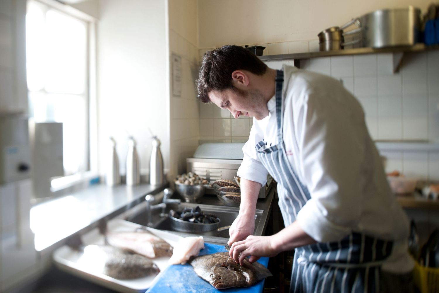 Chef preparing fresh fish on a cutting board in a professional kitchen, wearing a white chef coat and apron.
