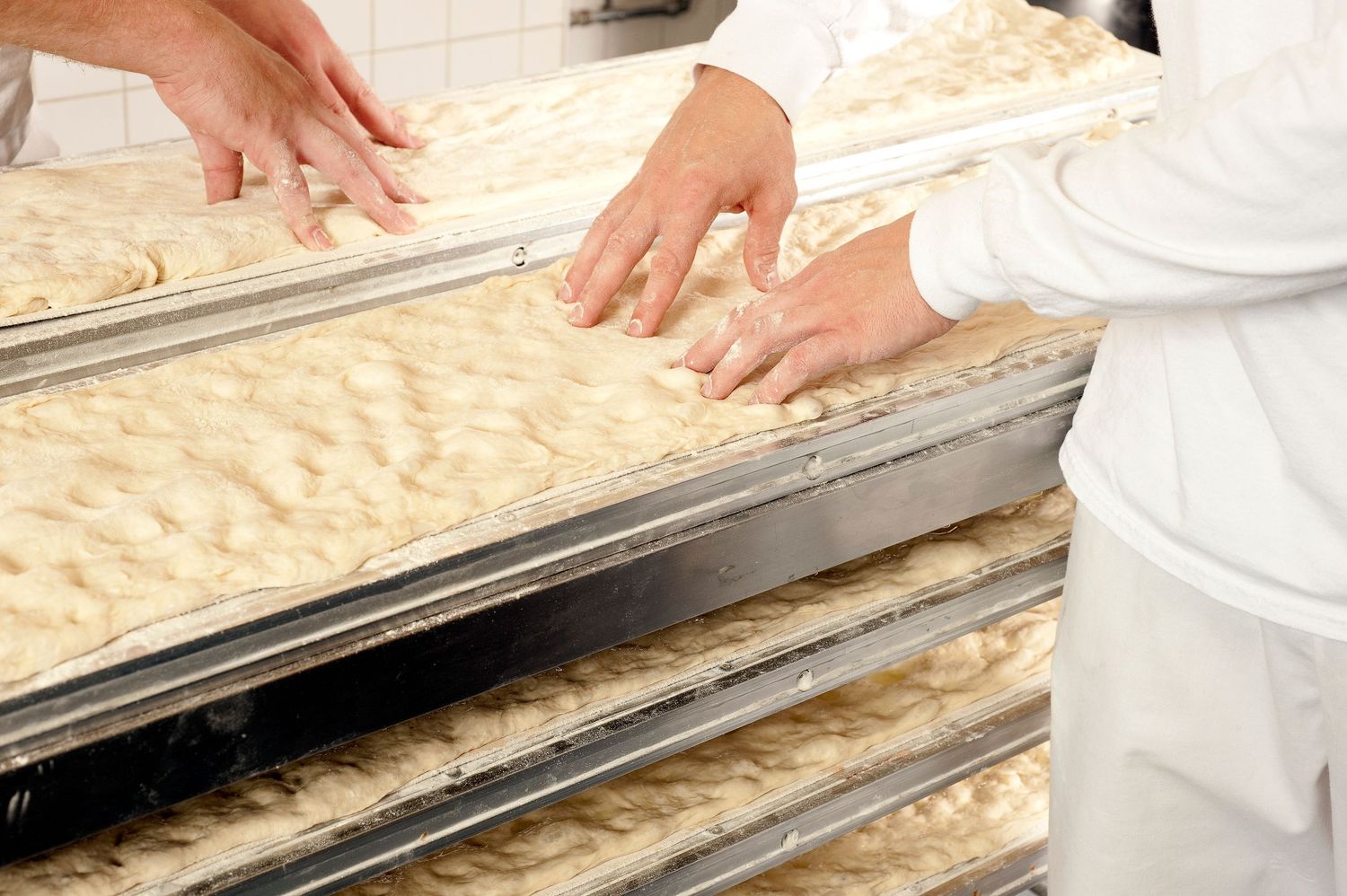 Bakers in white uniforms shaping raw dough on metal trays in a professional bakery production line.