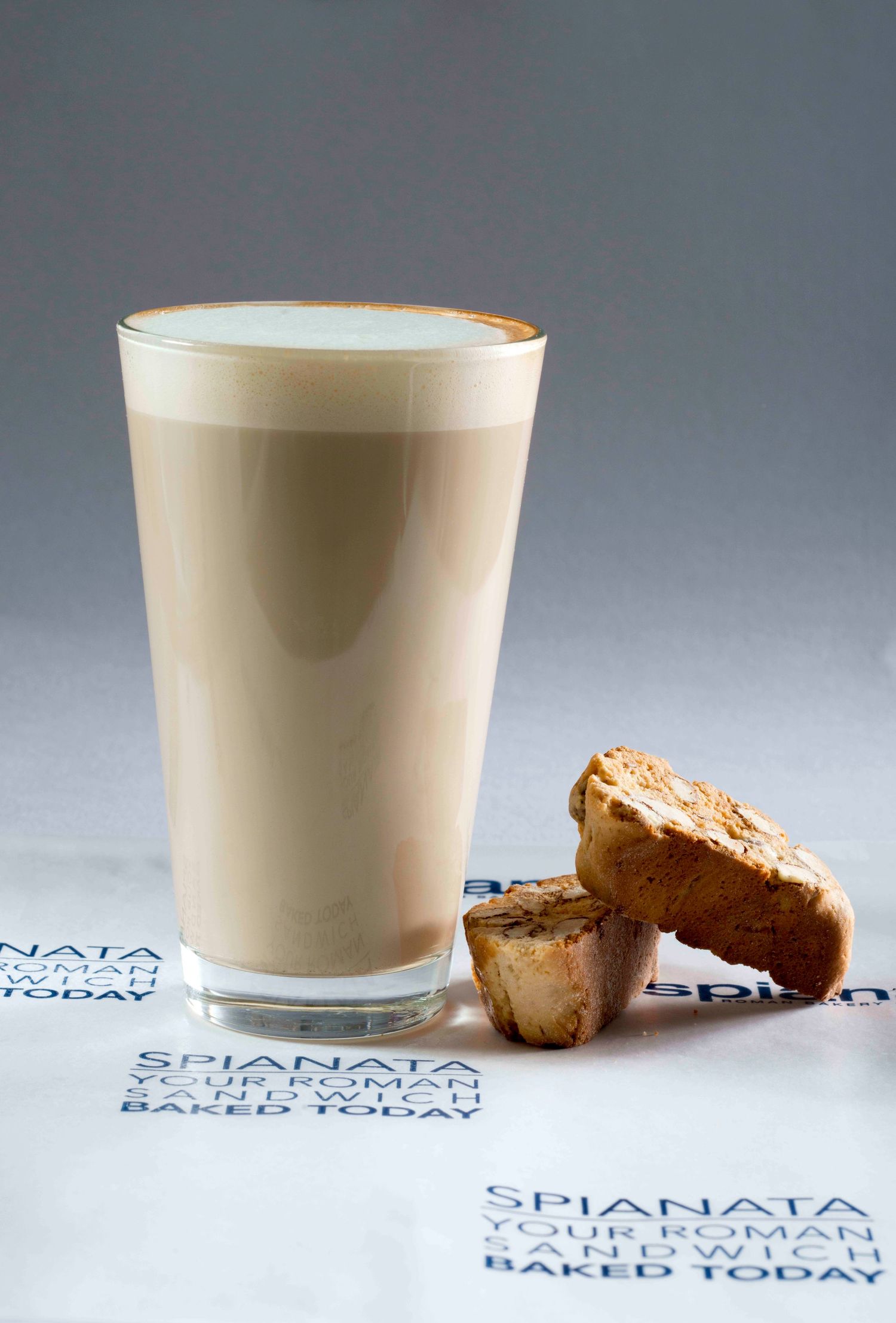 A tall latte glass with creamy coffee beside biscotti on a Spianata Roman sandwich branded napkin.
