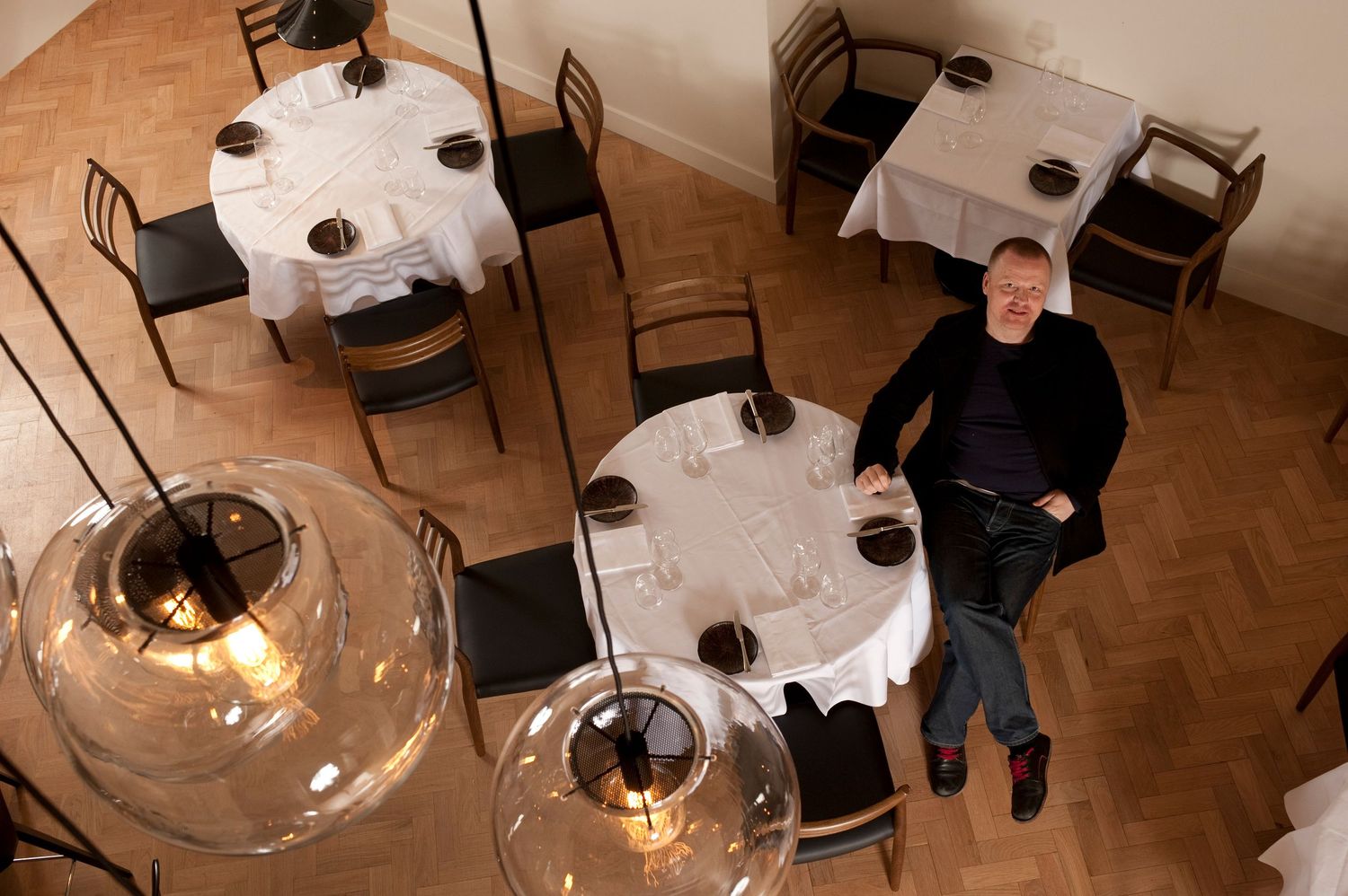 Man sitting alone at a white-clothed round table in an elegant restaurant with herringbone floors and pendant lights.