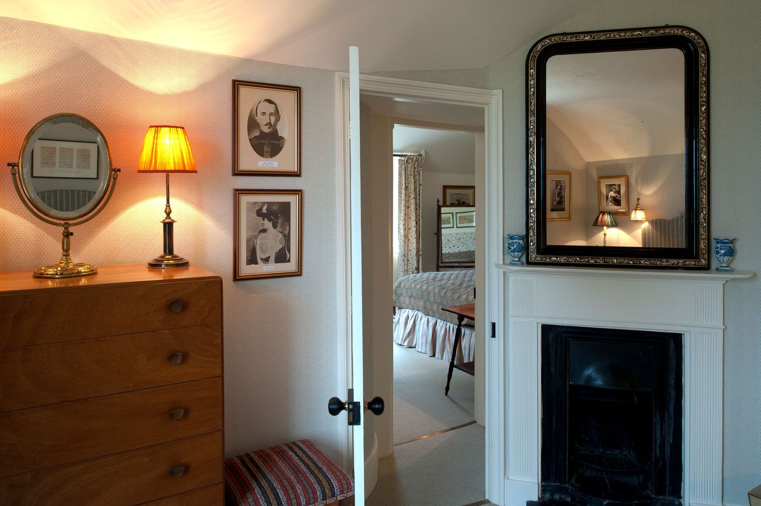 Cozy bedroom with wooden dresser, brass lamp, fireplace, ornate mirror, and doorway leading to an adjacent room.