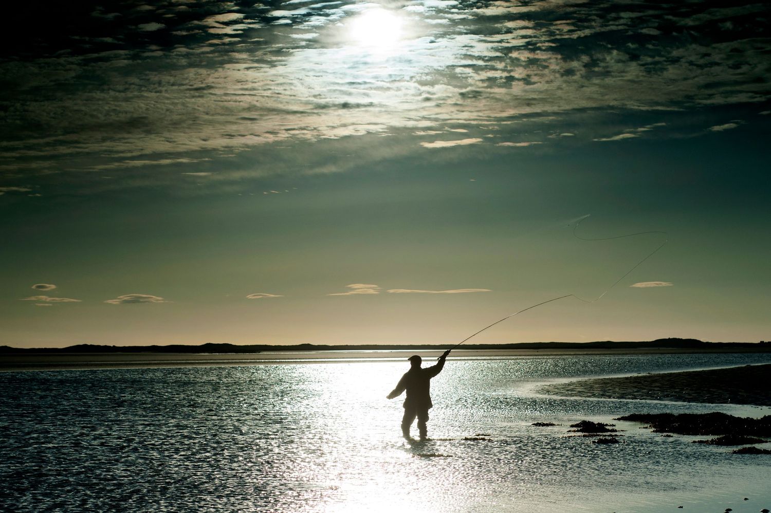 Silhouette of a fly fisherman casting a line in shallow water at sunset with dramatic cloudy sky.