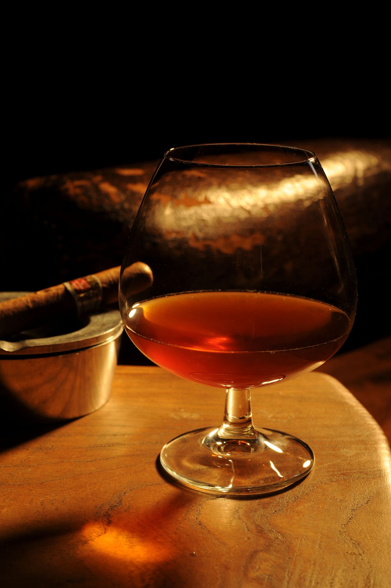 A snifter glass of amber cognac on a wooden table beside a cigar in dim, warm lighting.