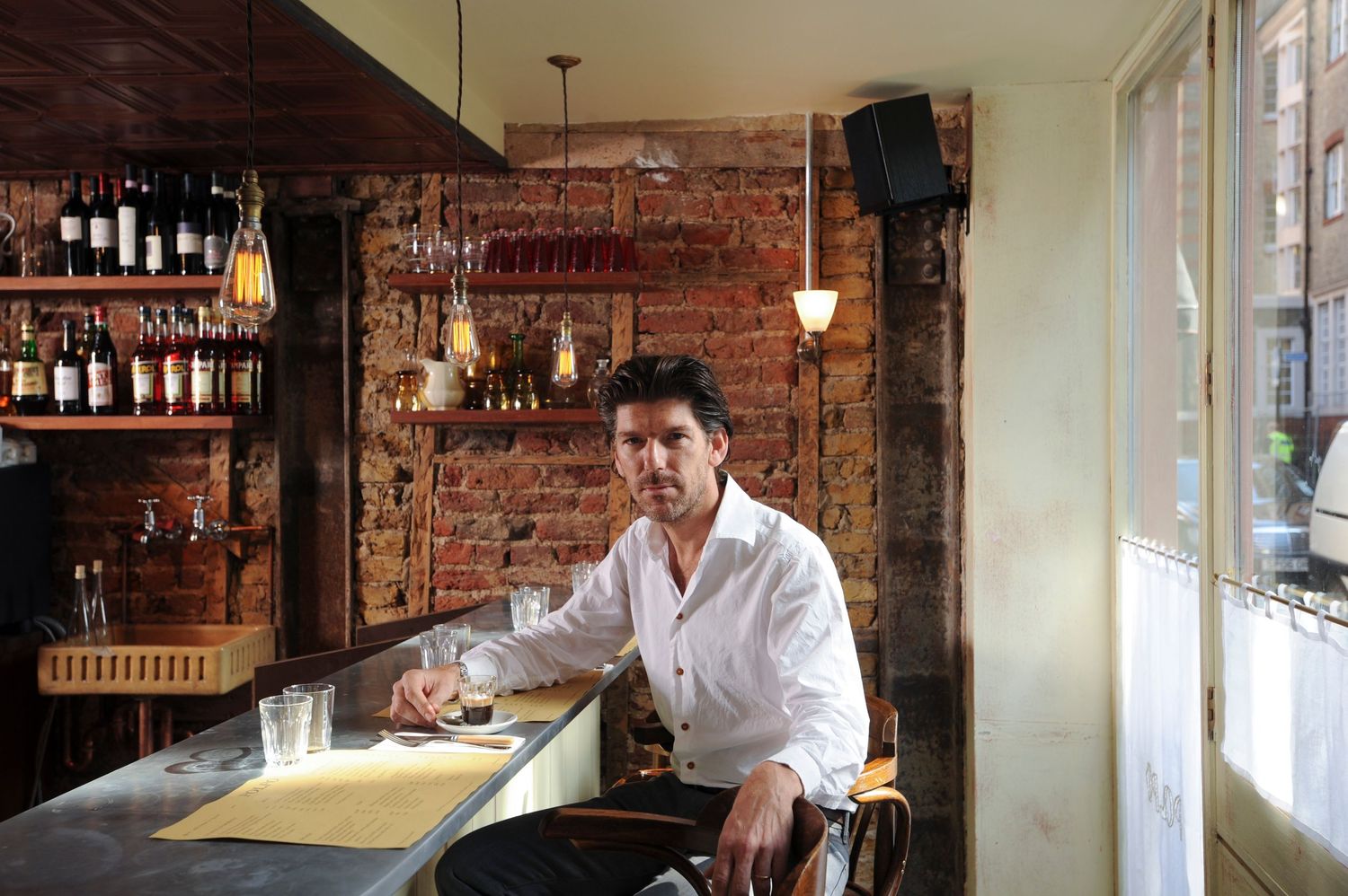 Man in white shirt sitting at a rustic bar with exposed brick walls and wine bottles on shelves.