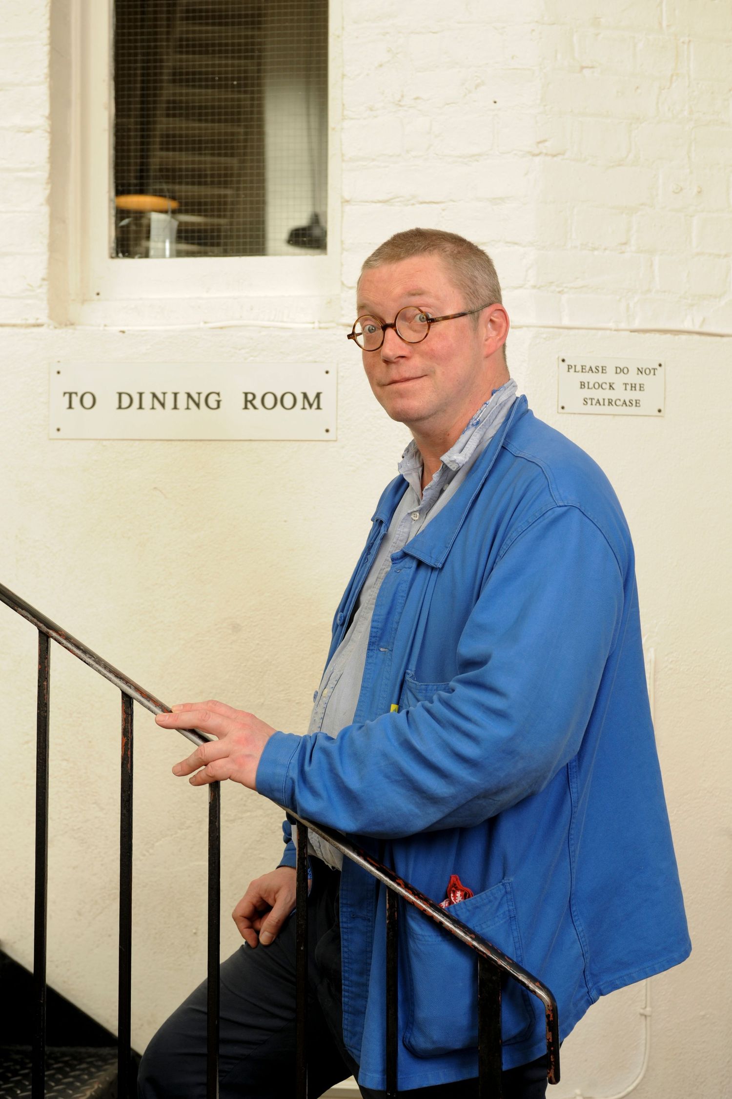 Middle-aged man in blue jacket holding stair railing near a 'To Dining Room' sign on white wall.