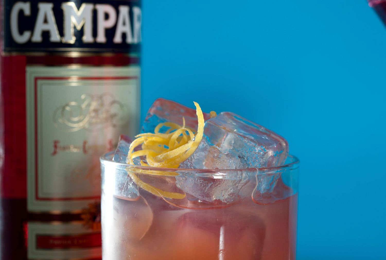 Campari cocktail on ice with lemon zest garnish in a glass, Campari bottle in background on blue surface.