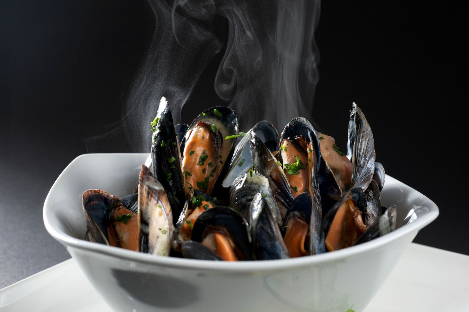 Steaming bowl of fresh cooked mussels in broth, served in a white ceramic bowl against a dark background.
