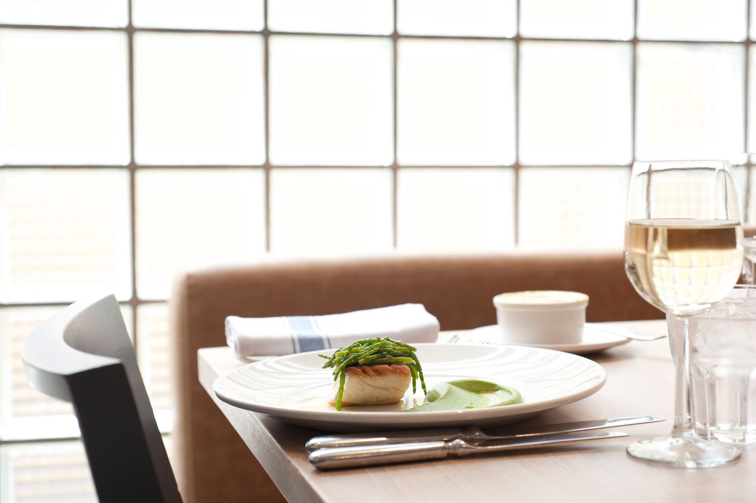 Elegant fine dining plate with gourmet dish, white wine glass, and modern restaurant setting by large windows.