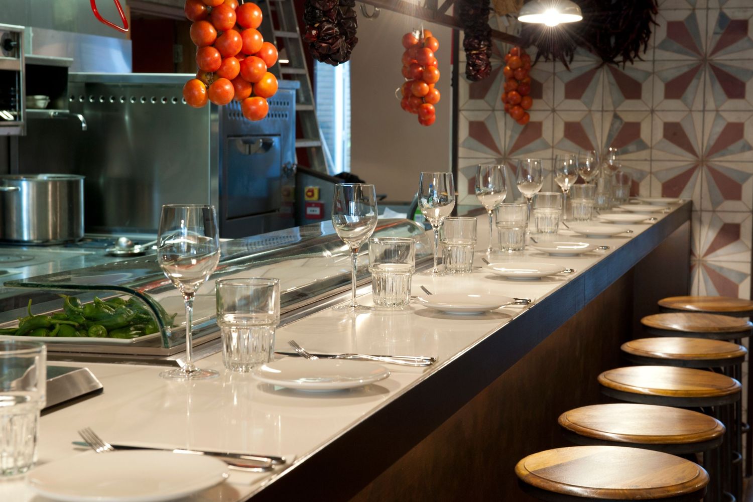 Elegant restaurant counter with wine glasses, white plates, and bar stools beside an open kitchen with fresh tomatoes.